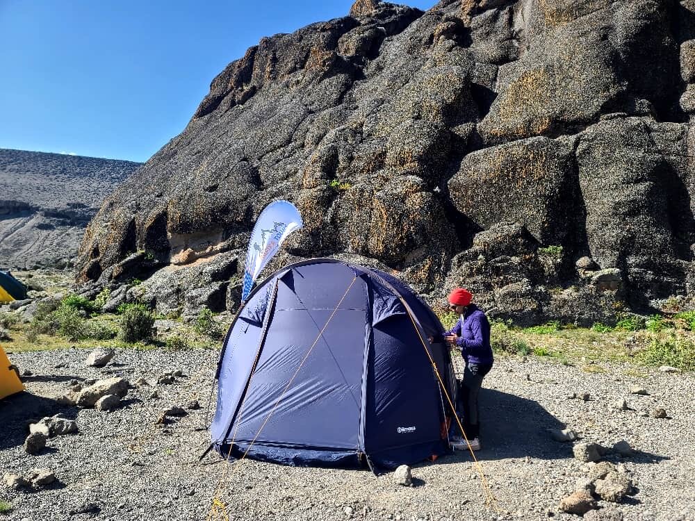 A trekker pitching a purple tent below a rock outcrop on Kilimanjaro